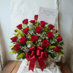 Canasta de mimbre con arreglo de rosas rojas, flores blancas pequeñas y follaje verde, decorada con lazo rojo y tarjeta sobre fondo claro