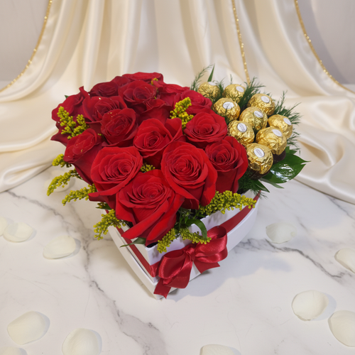 Caja en forma de corazón con rosas rojas frescas y chocolates dorados sobre mesa de mármol con pétalos blancos