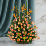 Arreglo floral grande con rosas naranjas y flores aves del paraíso sobre fondo de cortina verde y pared marmoleada