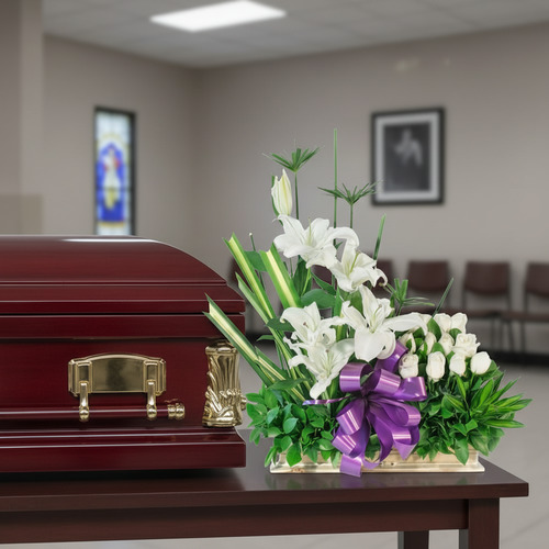 Arreglo de flores blancas con lazo morado sobre mesa junto a ataúd de madera barnizada en sala velatoria