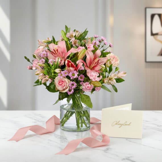 Arreglo floral ANNA con rosas rosadas, lirios y alstroemerias en jarrón de cristal, sobre mármol blanco con tarjeta de regalo
