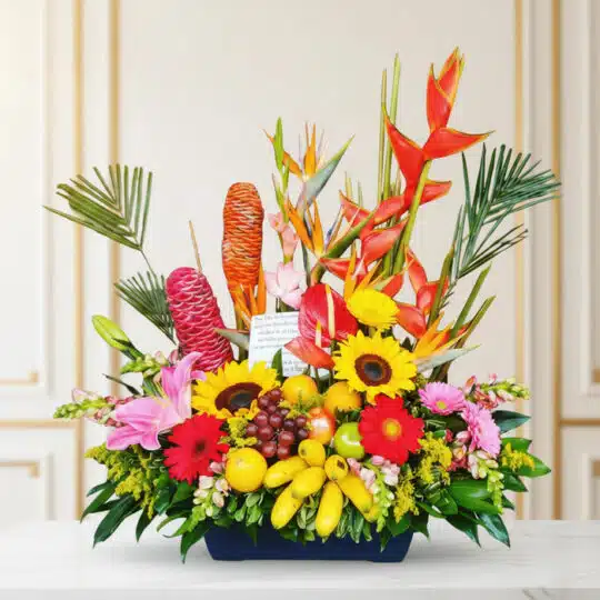 Arreglo floral exótico con heliconias, girasoles, lirios, anturios y frutas tropicales como plátanos, uvas, manzanas y limone