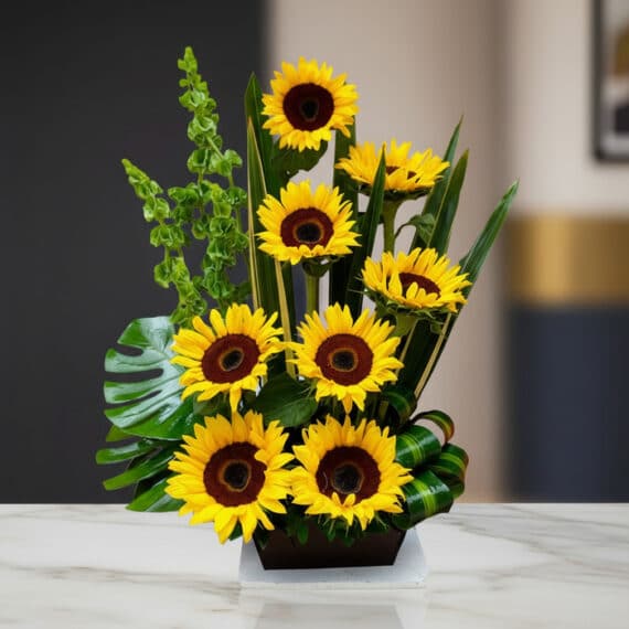 Arreglo Floral con Girasoles Bengala: Elegancia y Frescura para tu Hogar Arreglo floral premium con girasoles Bengala, hojas verdes y follaje, sobre una mesa de mármol en un lobby de hotel boutique.