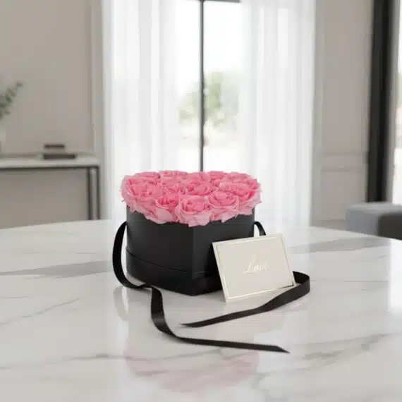 Arreglo Floral Perla con rosas rosadas en caja corazón negra, tarjeta de regalo 'Love' y cinta satinada sobre mesa de mármol.