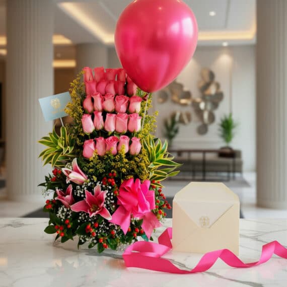 Arreglo floral elegante con rosas rosadas, lirios, follaje verde, globo fucsia, cinta de satén y sobre en mesa de mármol.