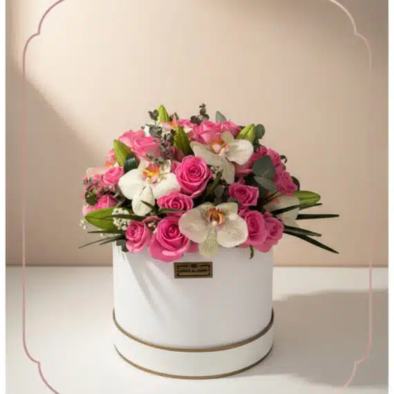Arreglo floral TANIA con rosas rosadas vibrantes y orquídeas blancas delicadas en una caja redonda blanca con borde dorado, i