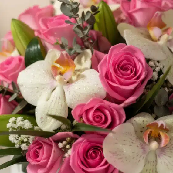 Primer plano extremo de un arreglo floral con rosas rosadas, orquídeas blancas con gotas de rocío, y follaje verde, destacand
