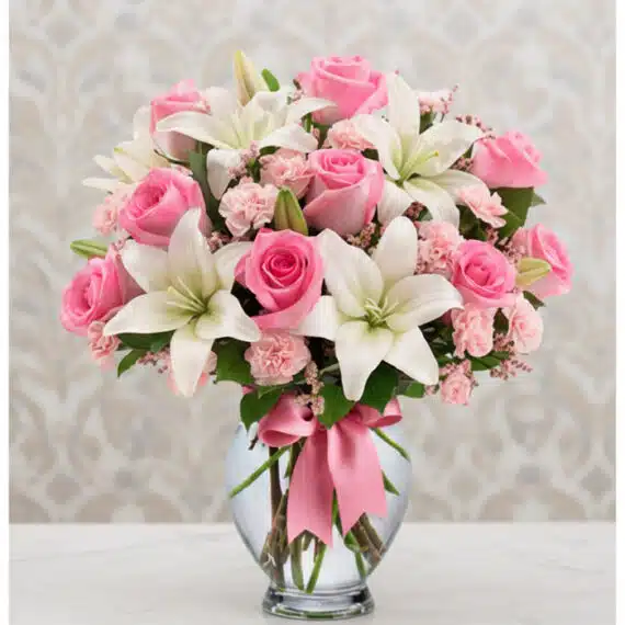 Arreglo Floral Violeta: Rosas Rosadas y Lirios Frescos en Cristal ✨ Arreglo floral vibrante con rosas rosadas, lirios blancos y claveles rosados en un jarrón de cristal con lazo rosa, ideal par