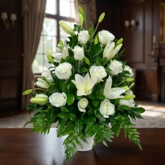 Arreglo Fúnebre Piedad: Rosas y Lirios Blancos Elegantes Arreglo fúnebre Piedad con rosas y lirios blancos, hojas verdes, en sala de velación premium con luz natural y bokeh.