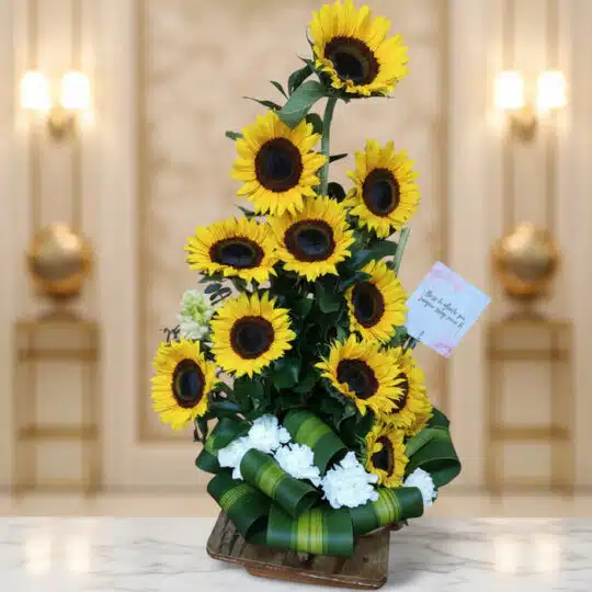 Arreglo floral de girasoles Sunna en un jarrón de madera, con detalles de flores blancas y lazo verde, sobre una superficie d