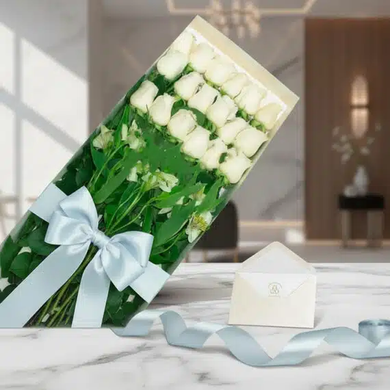 Caja de 18 rosas blancas frescas con follaje, lazo azul satinado, tarjeta de regalo y cinta decorativa sobre mármol pulido.