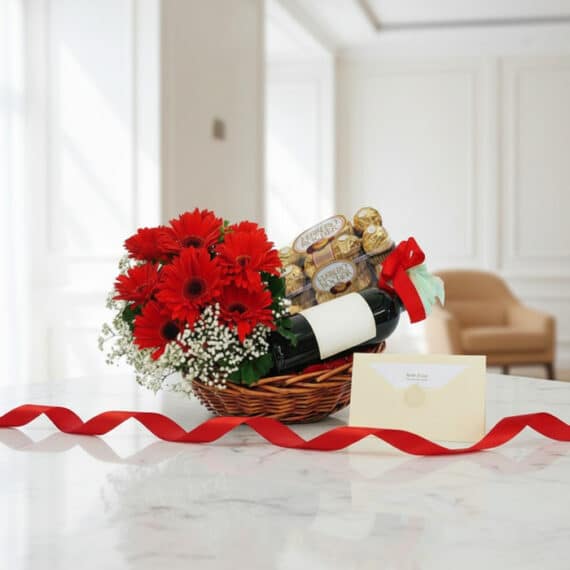Canasta Floral ROMA con gerberas rojas, vino y chocolates Ferrero Rocher, sobre mesa de mármol con tarjeta y cinta roja.