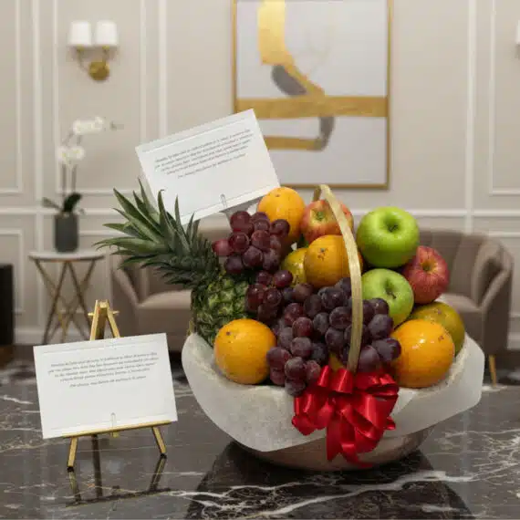 Canasta de Frutas Premium: Regalo Elegante y Saludable para Toda Ocasión Canasta de frutas premium con piña, uvas, manzanas rojas y verdes, naranjas, y un lazo rojo, sobre una mesa de mármol en un e