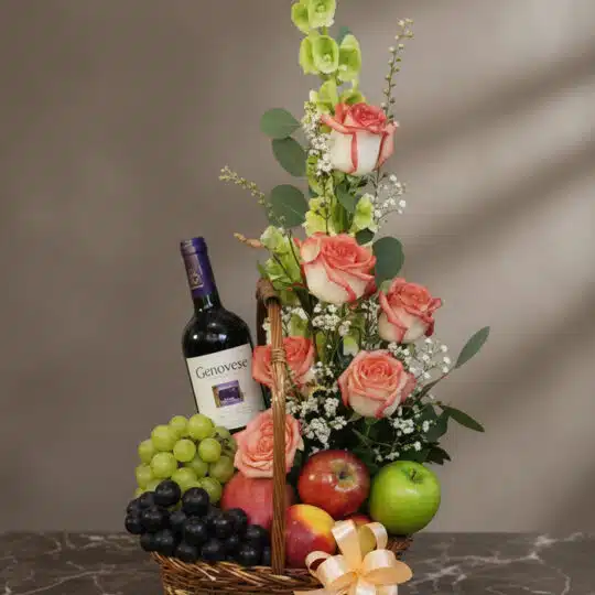Cesta gourmet CAPRICE con rosas frescas, uvas verdes y negras, manzanas rojas y verdes, duraznos y una botella de vino tinto