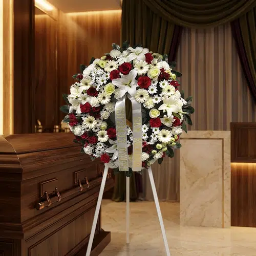 Corona de flores fúnebres Ararat con rosas rojas, lirios blancos y margaritas, junto a un ataúd de madera en un ambiente eleg