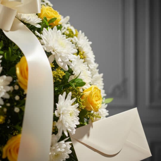 Primer plano de una corona fúnebre con rosas amarillas, crisantemos blancos y follaje verde, con una cinta satinada y un sobr