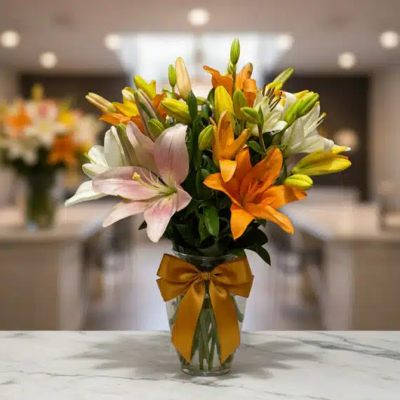 Jarrón con Lirios de Colores Vibrantes para Regalo Elegante Jarrón de cristal con un moño dorado, lleno de lirios de colores variados: blancos, naranjas, amarillos y rosas, sobre una su
