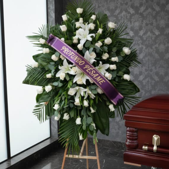 Pedestal fúnebre Eternidad con rosas blancas, lirios y follaje verde, adornado con una cinta morada que dice 'Profundo Pésame