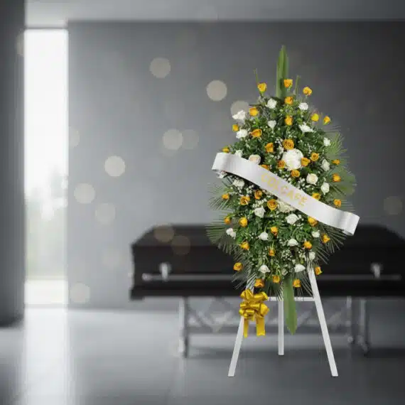 Pedestal fúnebre con rosas amarillas y blancas, follaje verde, y cinta blanca con el nombre 'COLCAFE', en una sala de velació