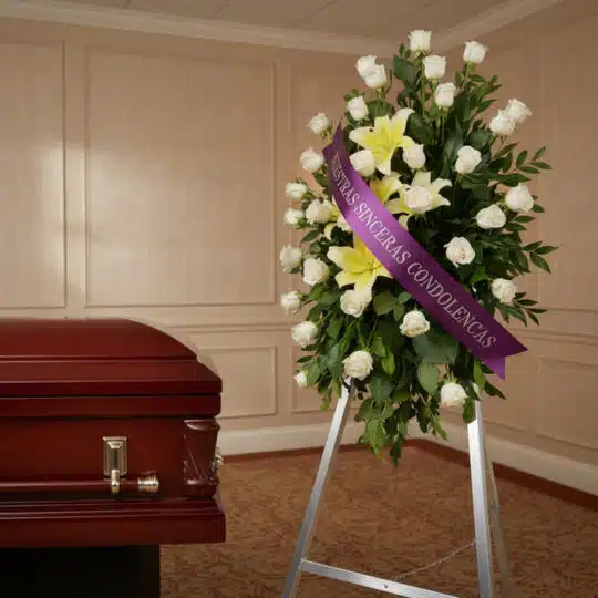 Pedestal fúnebre "Luz de Jacinta" con rosas blancas y lirios amarillos, cinta morada con mensaje de condolencia, junto a un a
