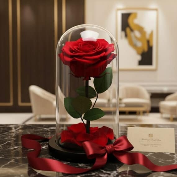 Rosa roja preservada en cúpula de cristal sobre mármol, con cinta de satén y tarjeta de regalo, ideal para un detalle sofisti